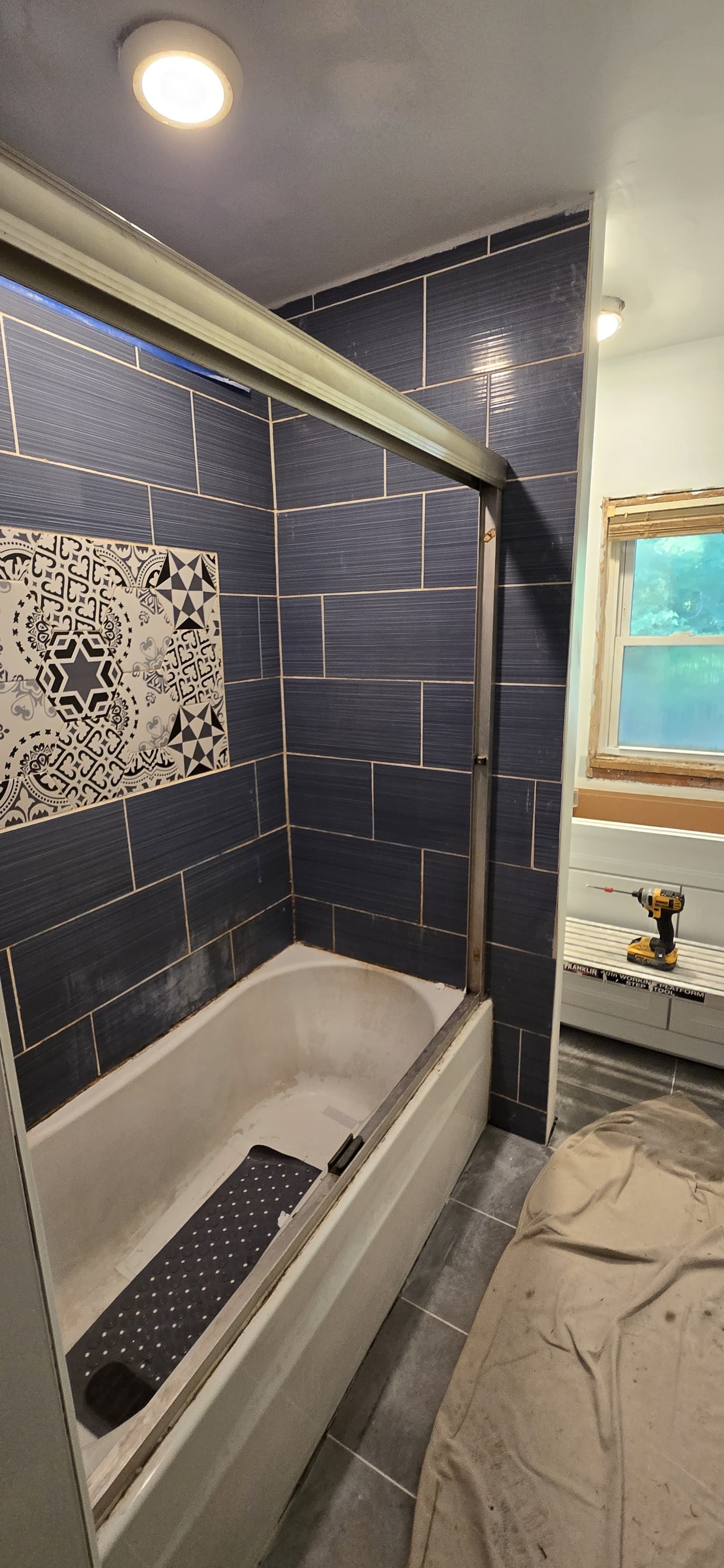 Bathroom shower enclosure with white soaking tub, dark blue subway tile walls with gold grout lines, decorative geometric tile accent, glass shower door, and ceiling light fixture