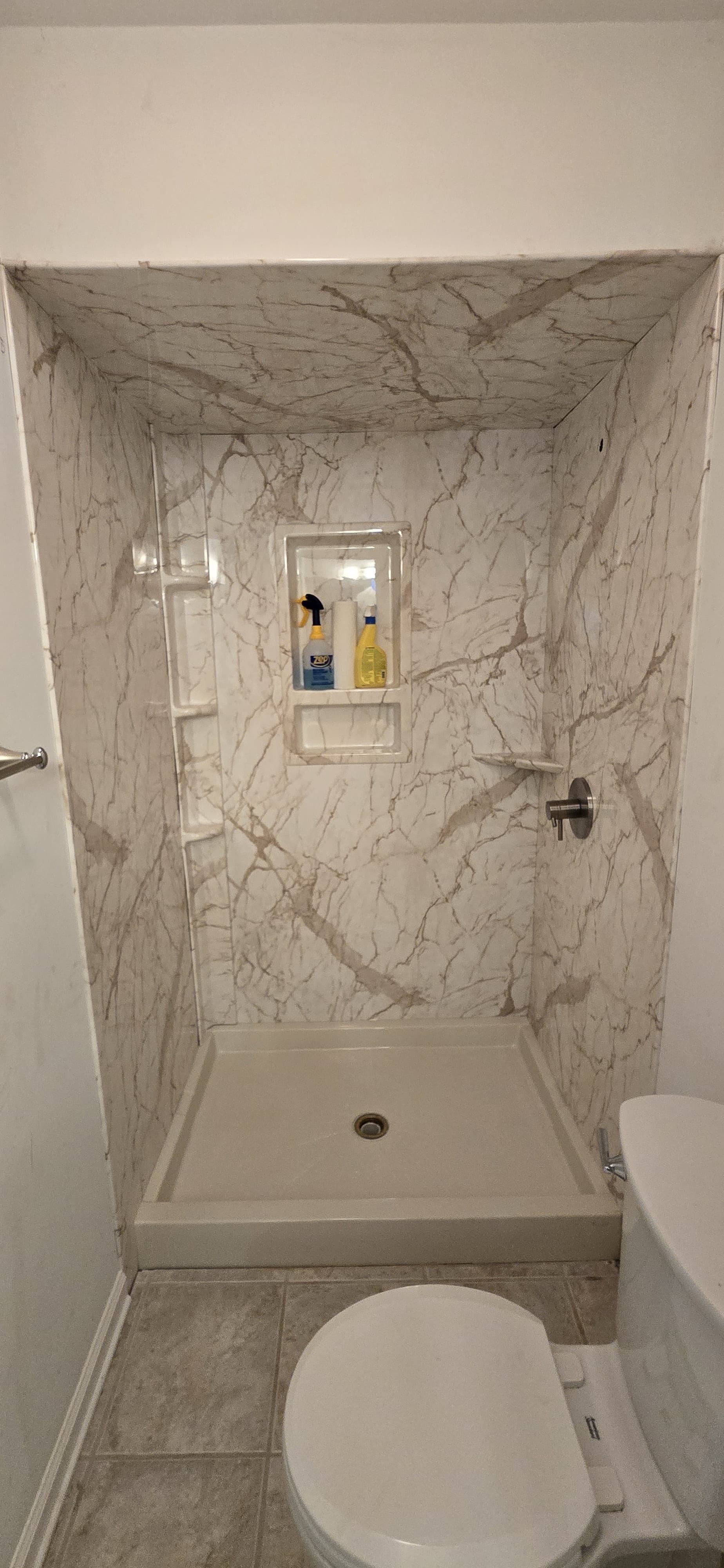 Marble-tiled shower enclosure with built-in shelf containing toiletries, adjacent to white toilet in bathroom