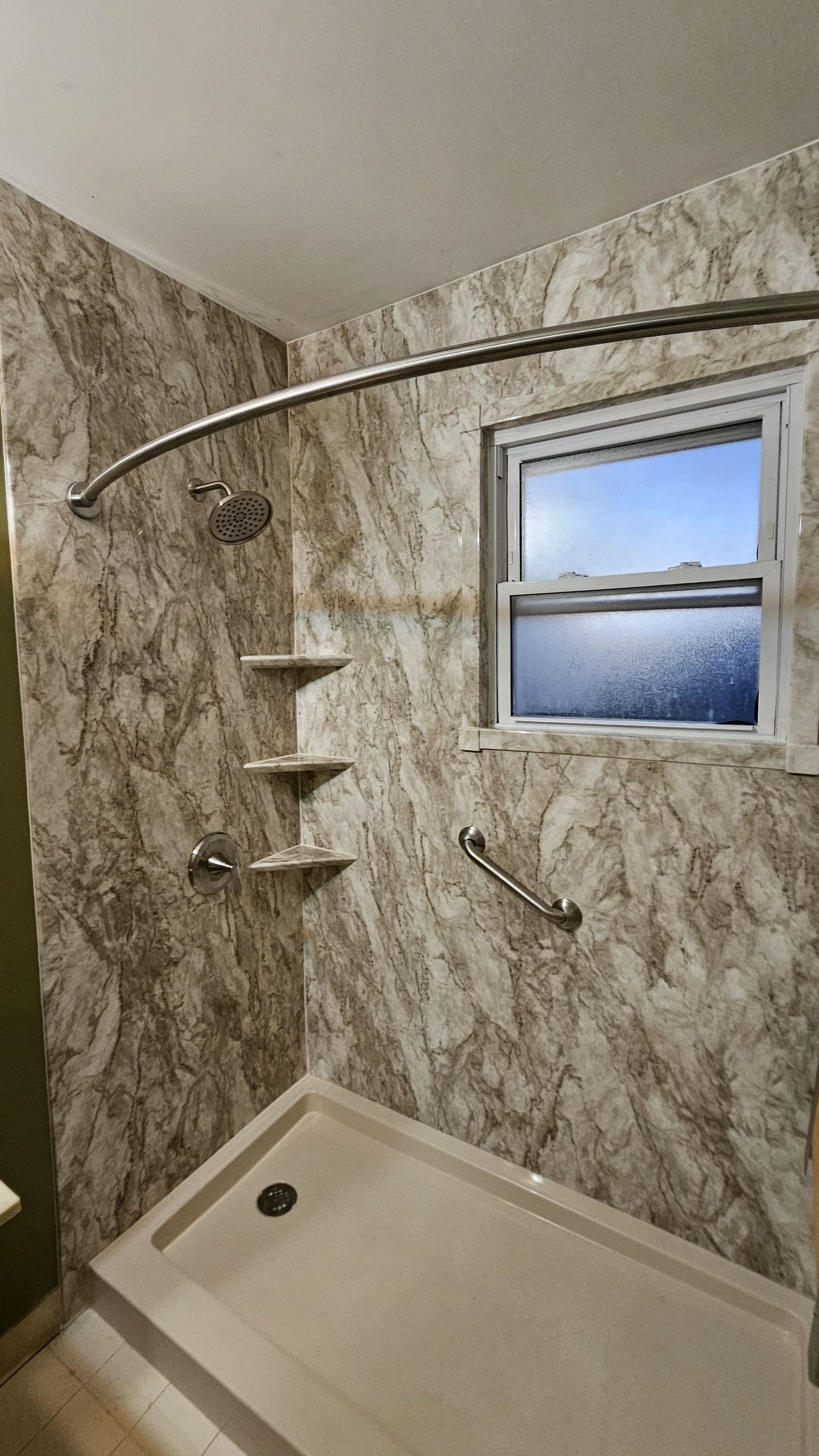 Modern shower stall with marble-look tile walls, curved chrome shower rod, showerhead, grab bars, built-in shelves, and a window with frosted lower pane