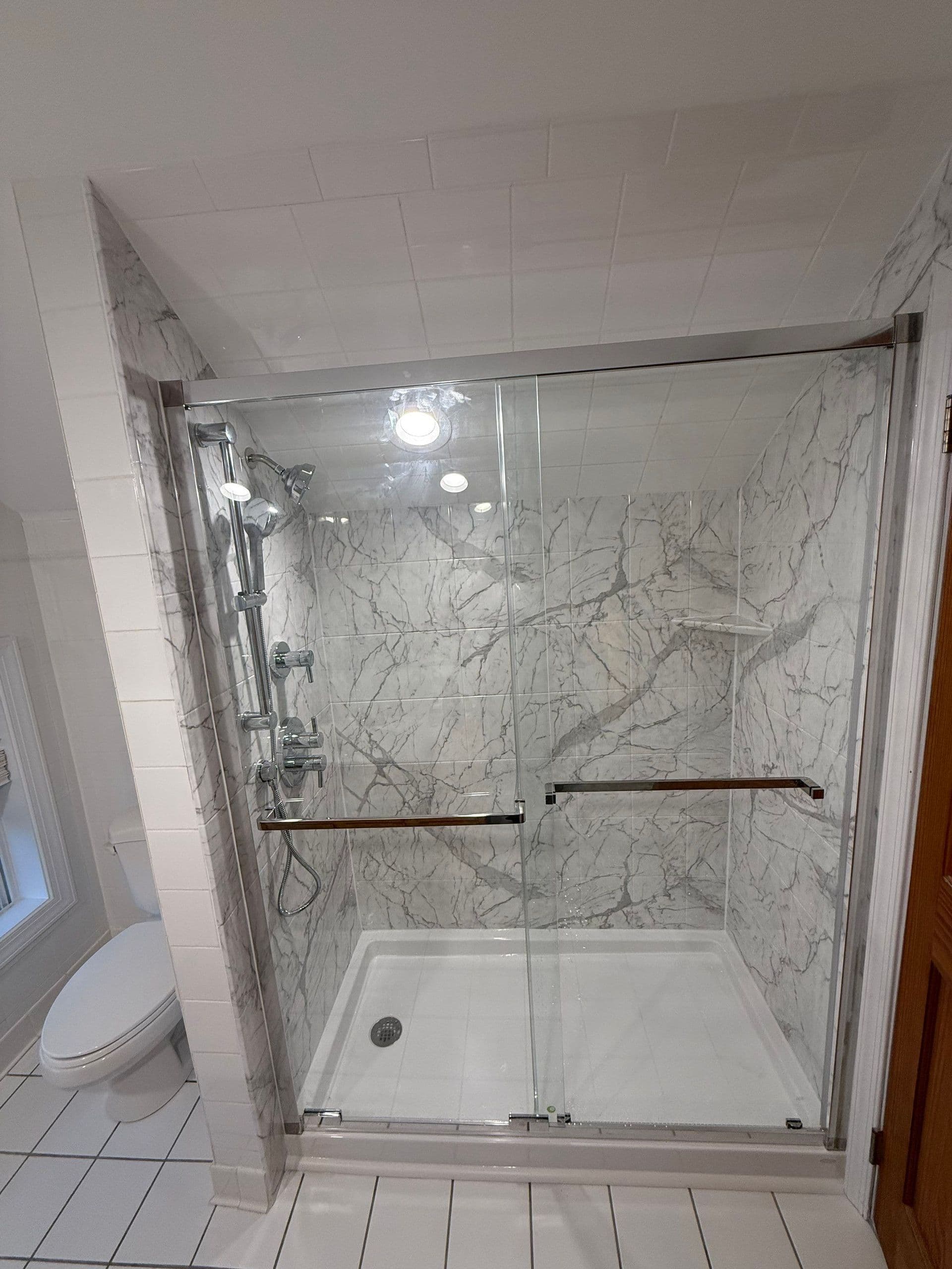 Glass-enclosed shower stall with marble-pattern tile walls, chrome fixtures, and rainfall showerhead in a bathroom with white subway tile walls and ceiling