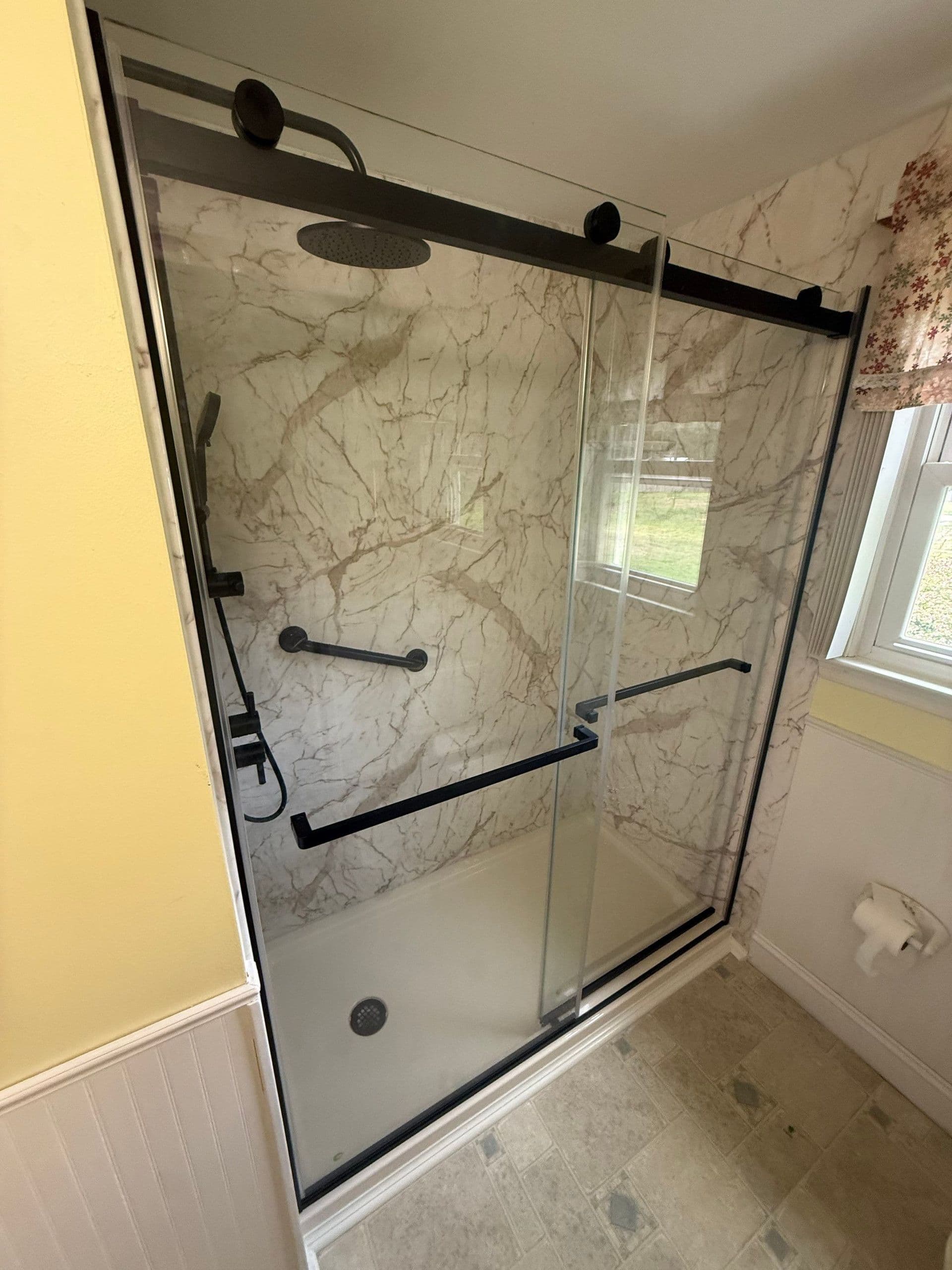 Frameless sliding glass shower enclosure with black hardware, marble tile surround, rainfall showerhead, and grab bars in modern bathroom