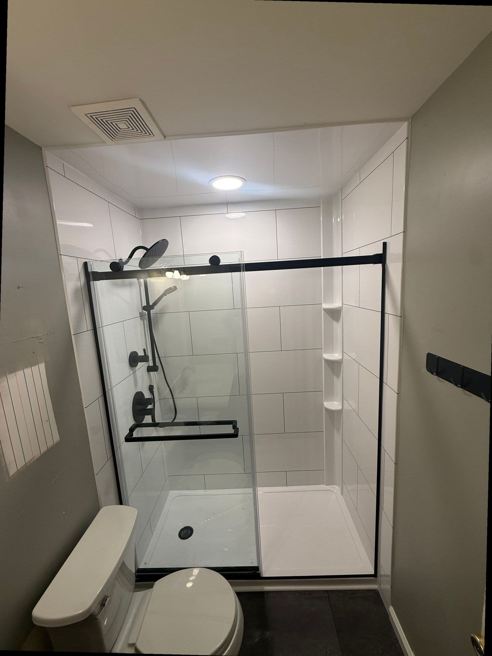 Modern bathroom with walk-in shower enclosure, rainfall showerhead, black fixtures, white tile walls, toilet, and built-in shelving