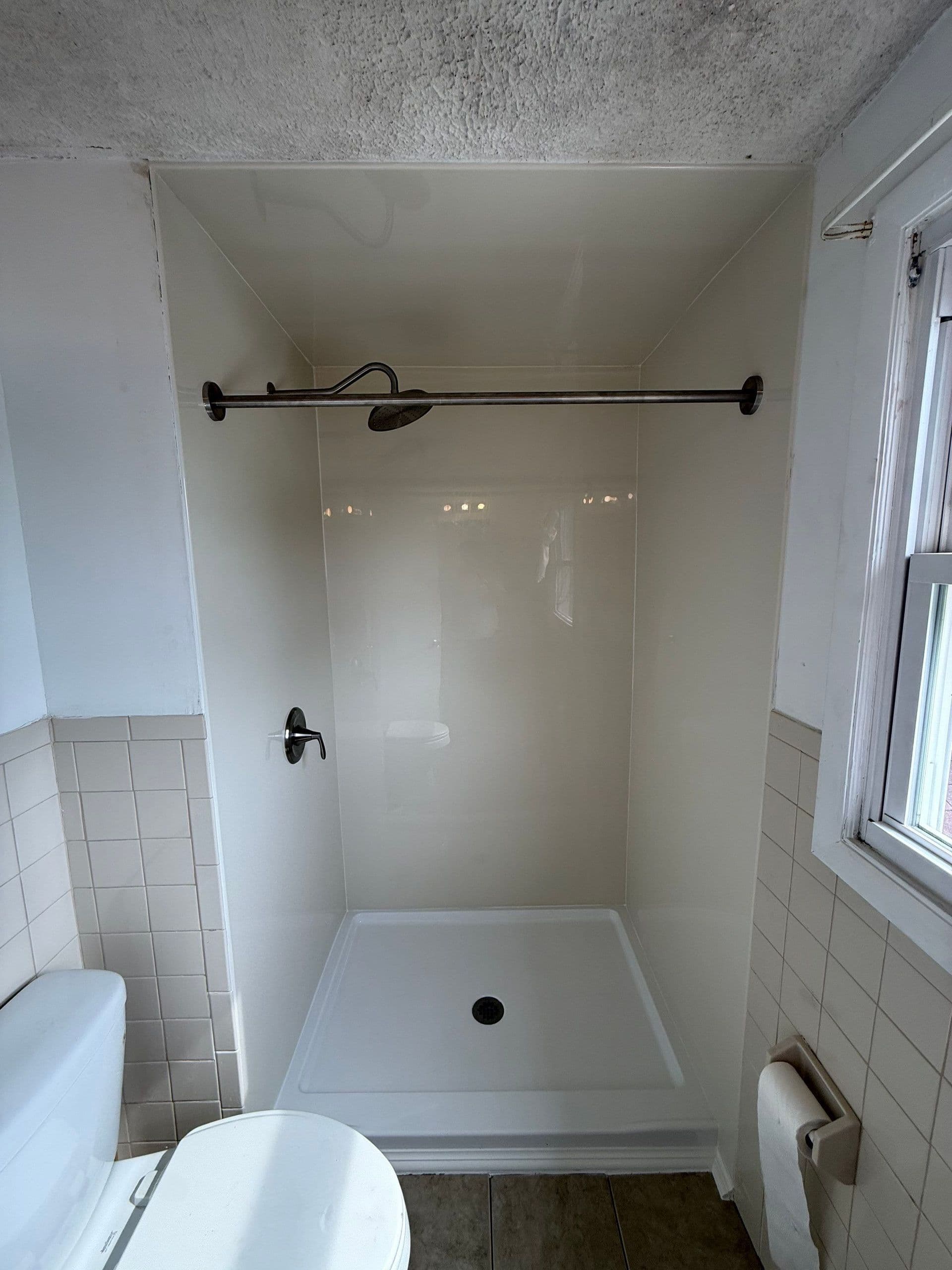 Bathroom shower enclosure with white fiberglass walls, black showerhead and controls, white base pan, and sliding glass door on track mounted above