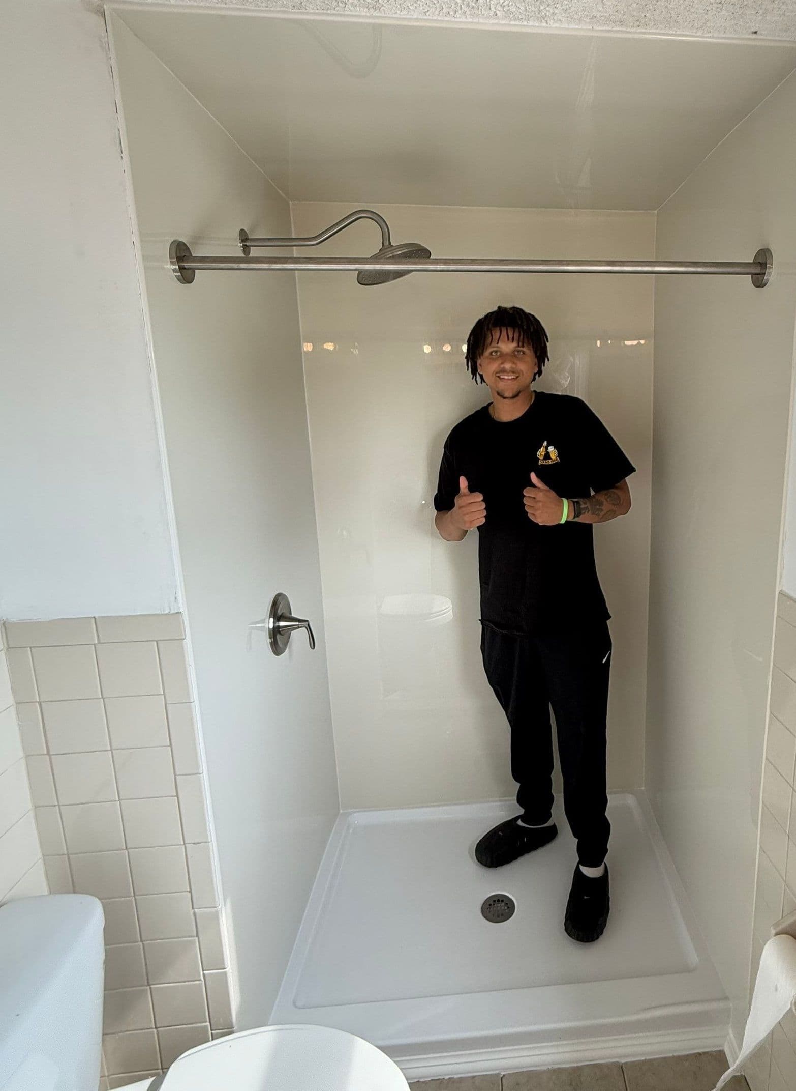 Person giving thumbs up while standing in a newly renovated shower stall with white tile walls and metal showerhead