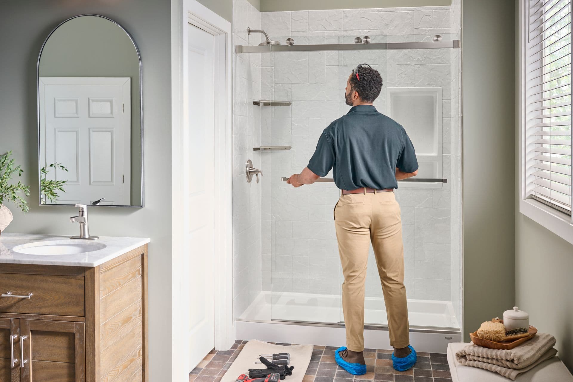 A worker installs sliding glass doors in a gray bathroom with a white tiled walk-in shower.