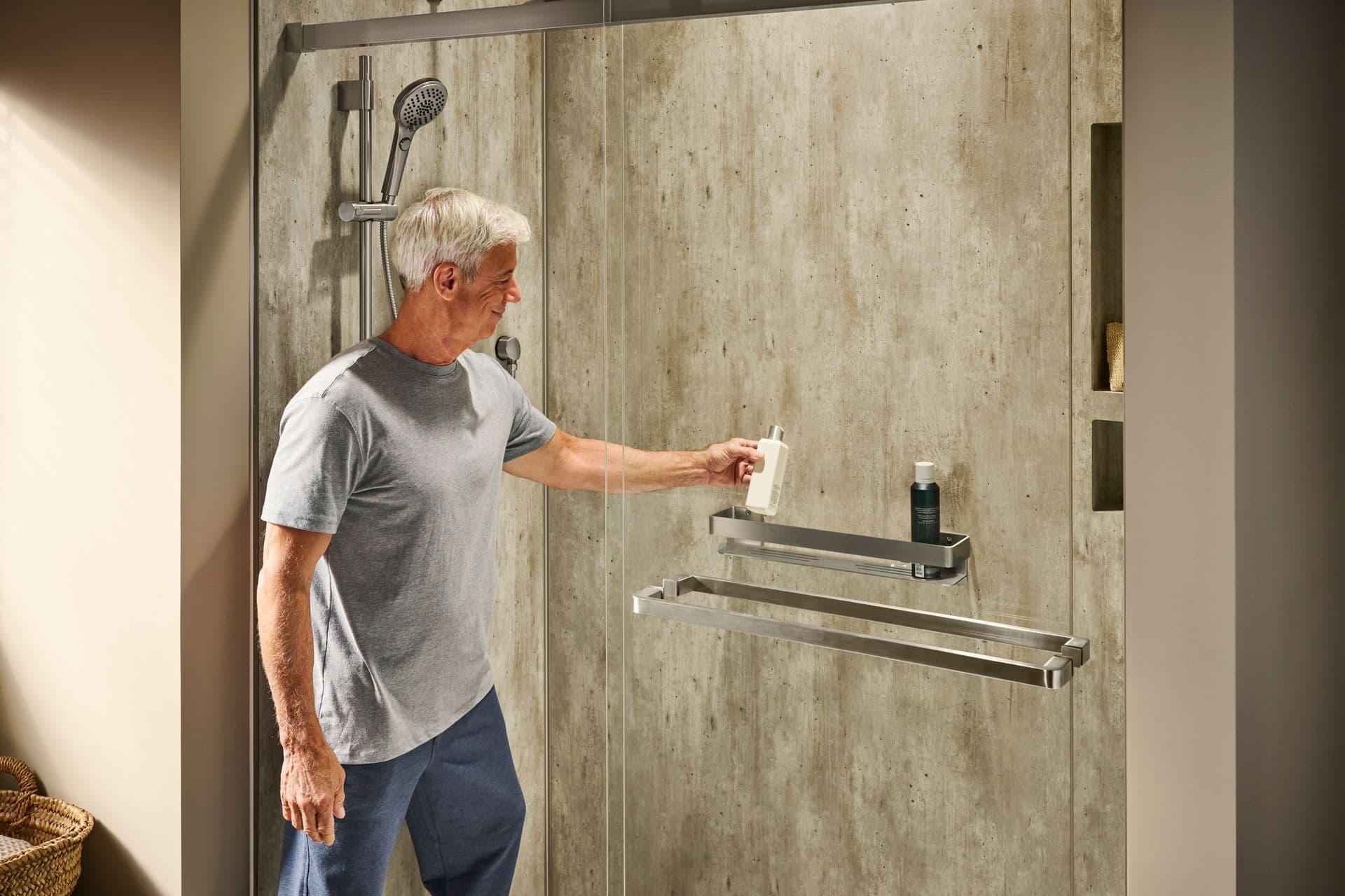 A person places a bottle on a custom shelf in a walk-in shower with gray textured walls, sliding glass doors, and brushed steel fixtures in a tan bathroom.