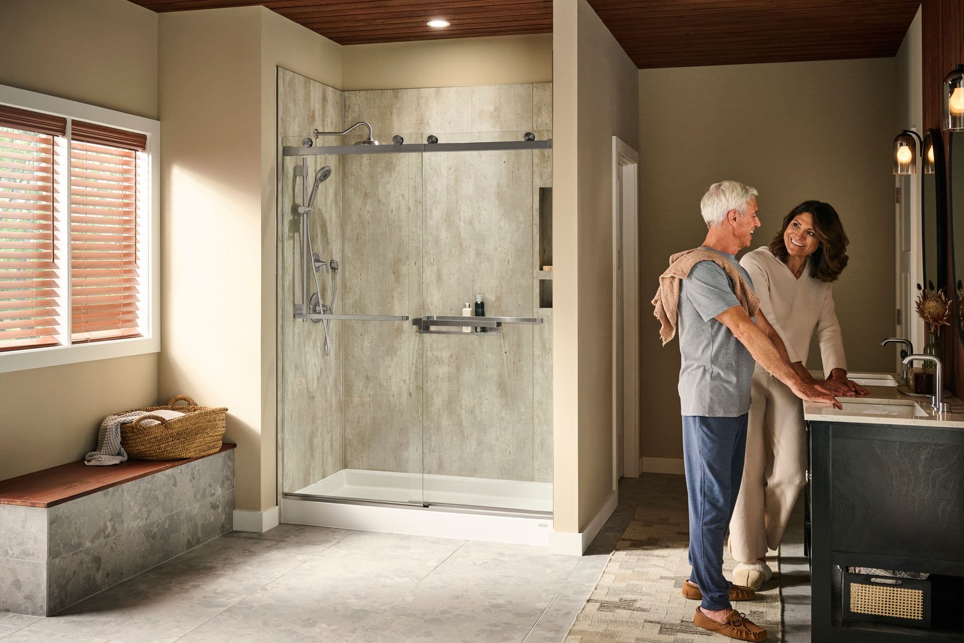 A couple stands in front of a double sink in a beige bathroom with a walk-in shower with gray textured walls, sliding glass doors, and brushed steel fixtures.
