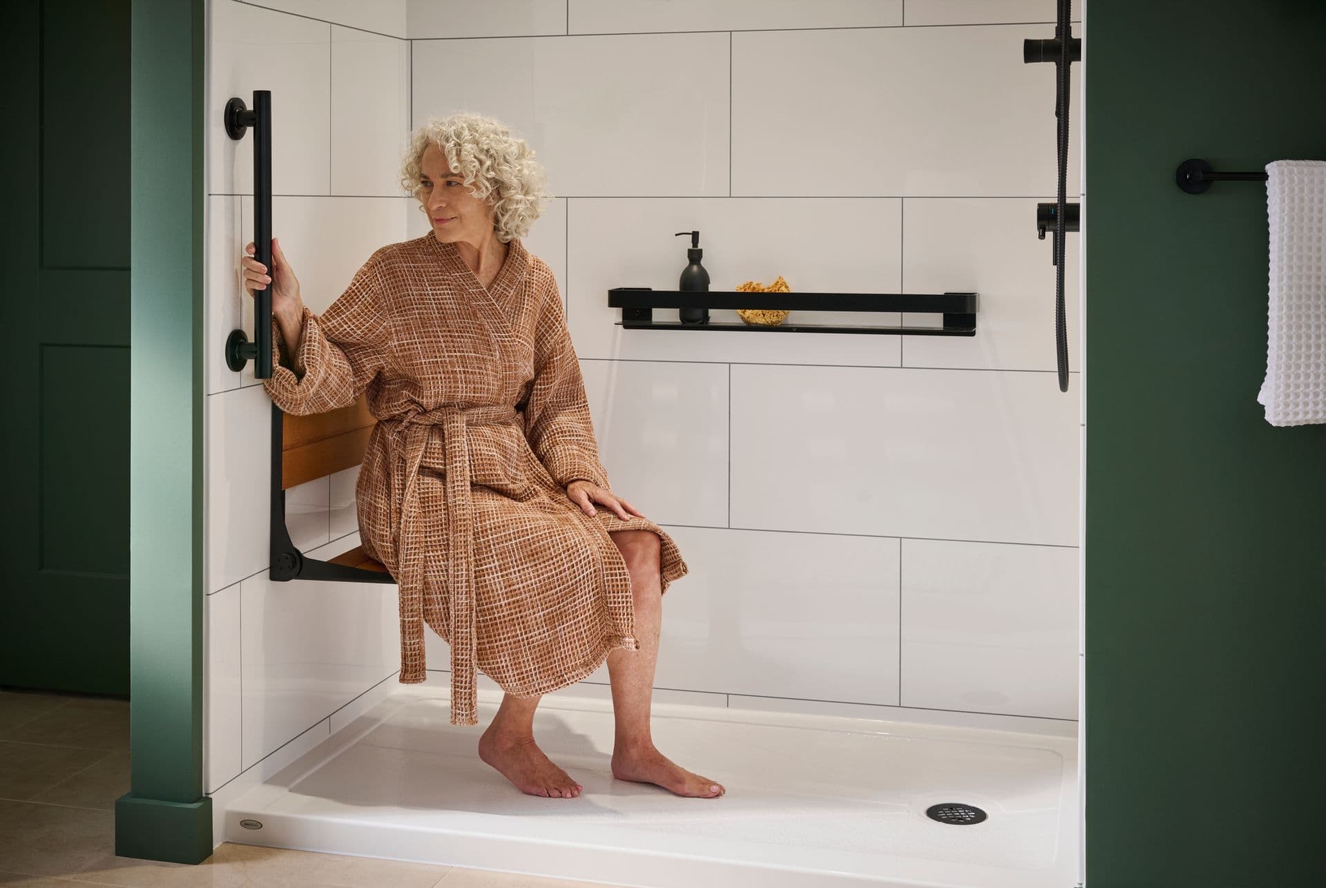 A person sits on a built-in shower bench and holds onto a black vertical grab bar in a shower with large white tiled walls in a green bathroom.