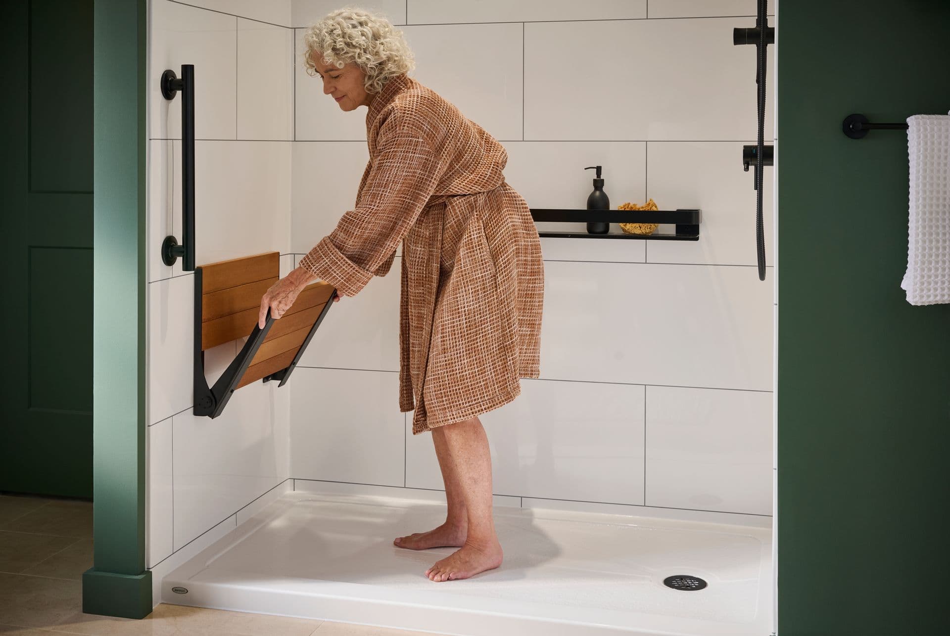 A person unfolds a built-in shower bench in a green bathroom with white tile shower walls and black fixtures.