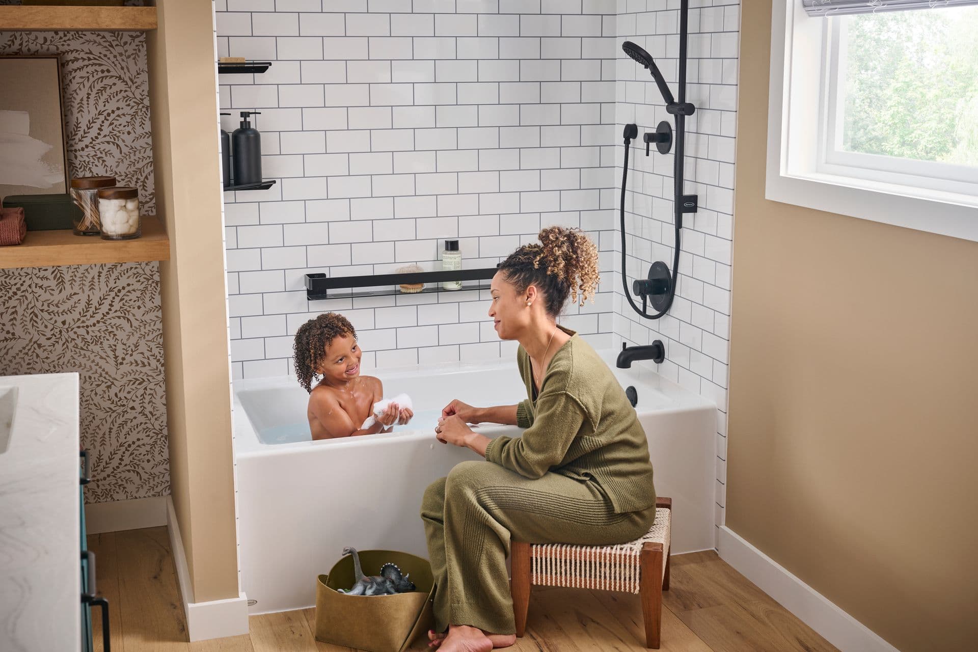 A parent sits next to a child in a bathtub with white tile walls and black fixtures in a tan bathroom.