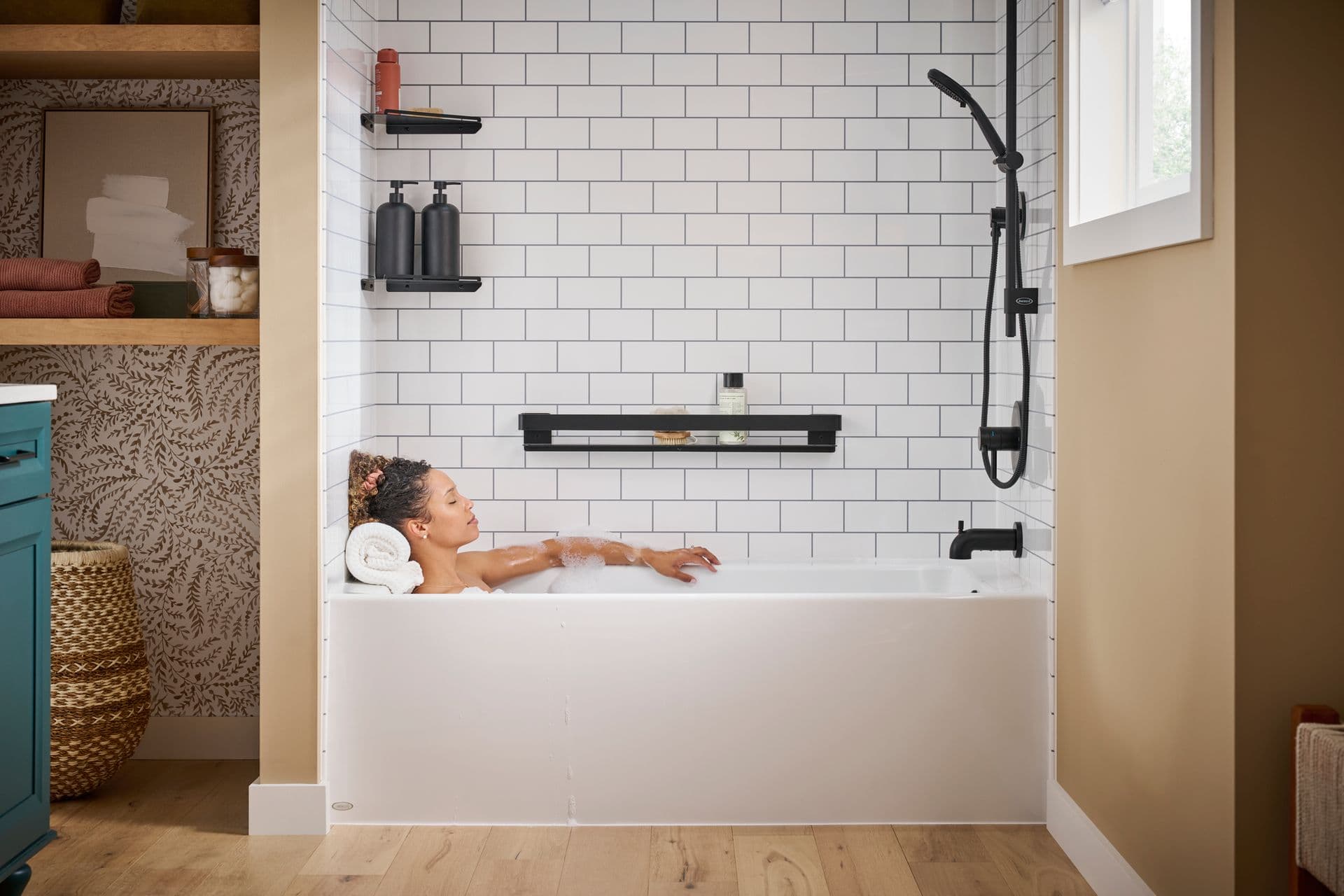 A person relaxes in a combination bathtub shower with white tile walls and black fixtures in a tan bathroom.