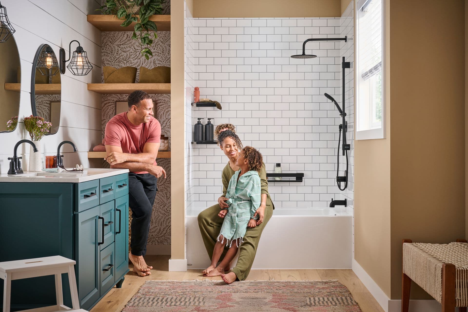 A family in a tan bathroom. A parent and child sit on the edge of a combination shower bathtub with white tile walls and black fixtures.